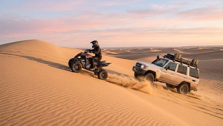 Quad bike following a Land Cruiser down a rippled dune in soft sunrise light